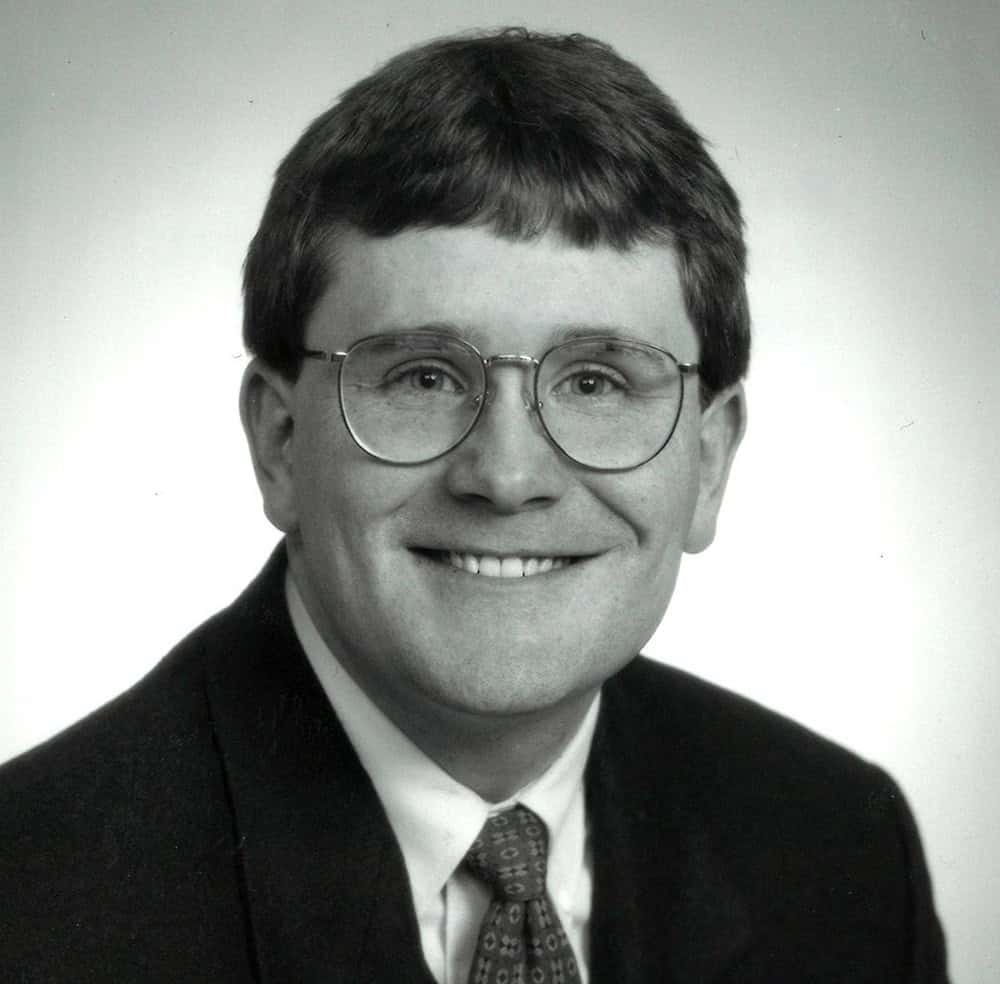 Black and white portrait of a smiling man in glasses and suit.