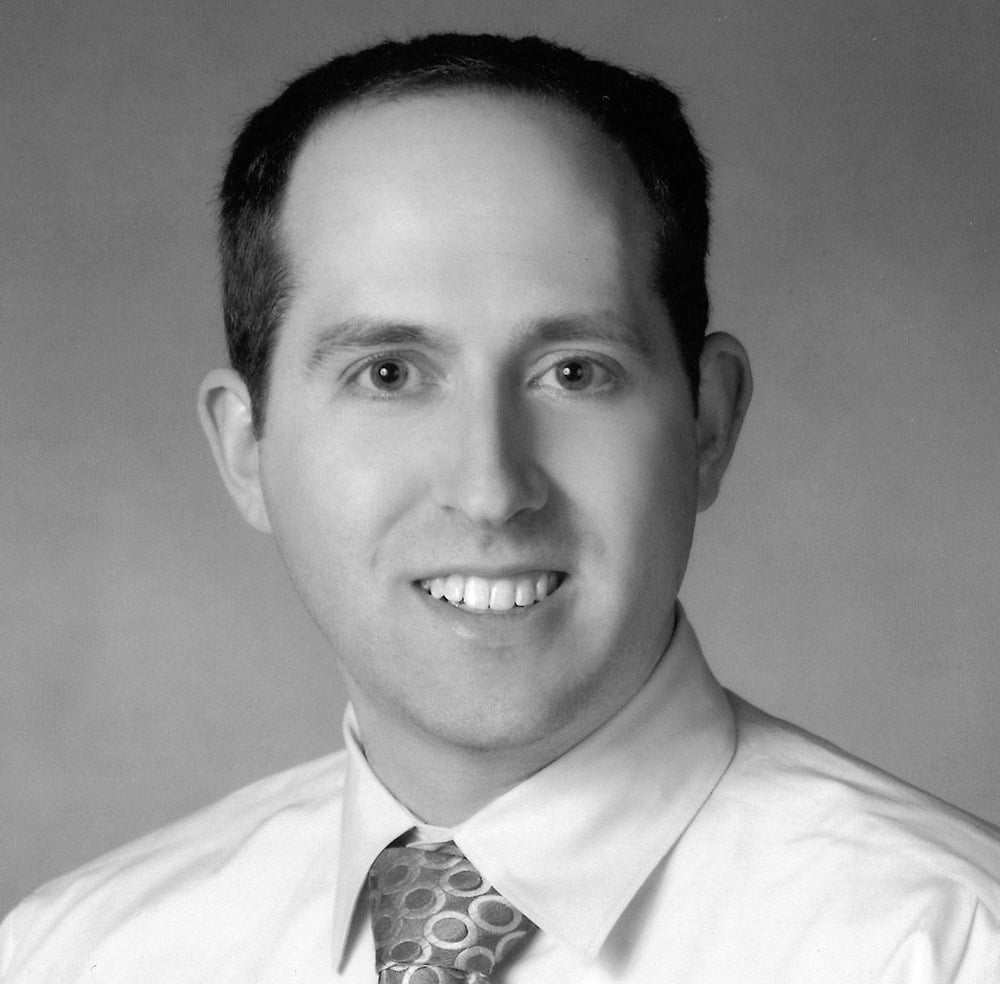 Professional portrait of a man in a white shirt and patterned tie.