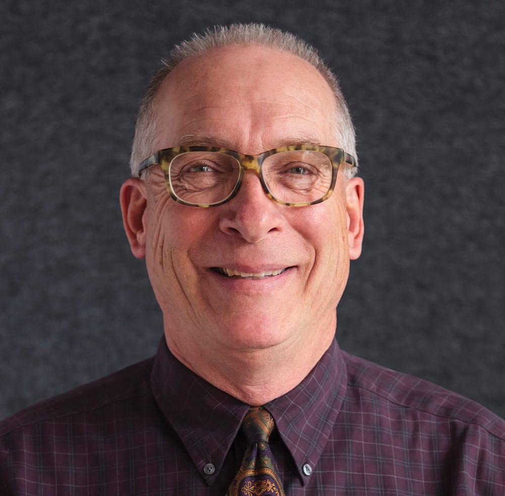 Smiling man with glasses and gray hair, wearing a checkered shirt and tie.